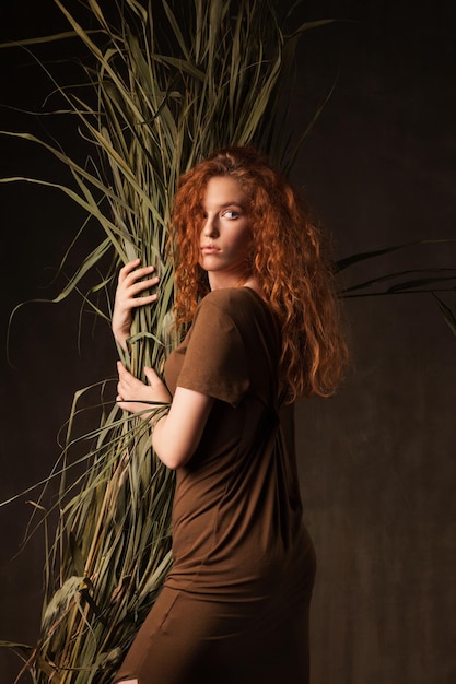 Energizer piękna młoda kobieta z kręconymi, rudymi włosami z zieloną tropikalną trawą z trzciny cukrowej postawiona w ciemnych ścianach studio Kopiuj tło