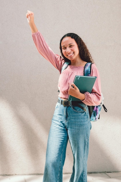 Entuzjastyczna czarna studentka podnosząca rękę i trzymająca książki przy ścianie. Edukacja