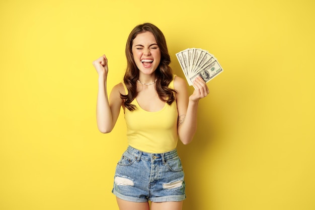 Entuzjastyczna uśmiechnięta kobieta pokazująca gotówkę, śmiejąca się i wyglądająca na podekscytowaną, stojąca w letnich ubraniach na żółtym tle