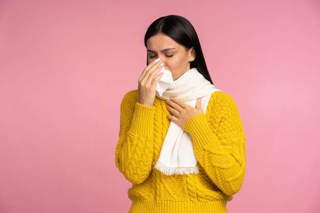 Epidemia wirusa, epidemia grypy. Chora kobieta owinięta chustą, kichająca w serwetkę, oczyszczająca z nosa i kaszląca, cierpiąca na objawy grypy, gorączka. "Kryty studio strzał na białym tle na różowym tle"