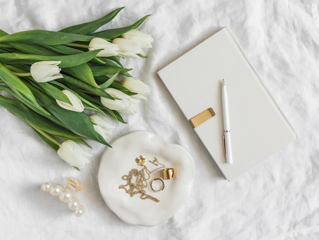 Estetyczne akcesoria dla kobiet biżuteria na ceramicznym talerzu notatnik bukiet białych tulipanów na jasnym tle widok z góry