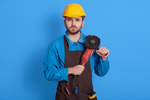 Europejski mężczyzna robotnik budowlany z szlifierką w rękach pozowanie na białym tle nad niebieską ścianą, patrzy na aparat z poważną miną, ubrany w ochronny kask i fartuch, brygadzista jest gotowy do pracy.