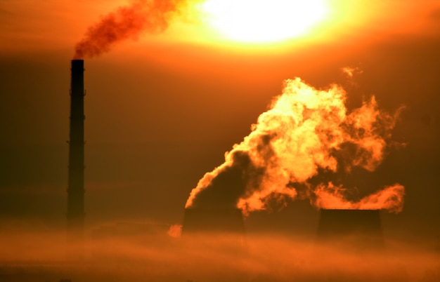 Zdjęcie fabryka z kominami i dymem w pomarańczowym blasku świtu i porannej mgle w lecie. jasne słońce świeci przez elektrownię dymu, elektrownię. tło przemysłowe