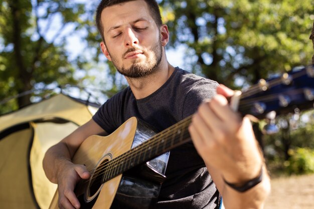 Facet gra muzykę na gitarze w pobliżu namiotu na składanym krześle w lesie