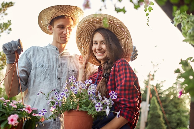 Facet i dziewczyna ogrodnicy w słomkowych kapeluszach trzymają na ogrodowej ścieżce w słoneczny dzień doniczki ze wspaniałymi petuniami. .