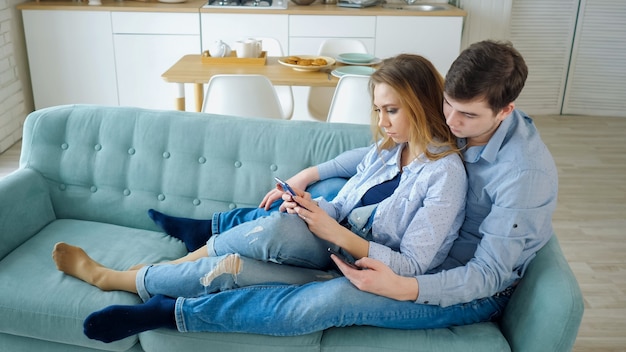 Facet i dziewczyna siedzą na kanapie surfując po Internecie w smartfonach, widok z boku