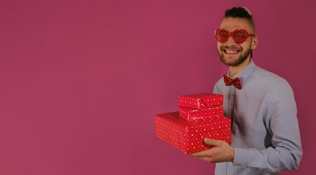Facet nosi okulary w kształcie serca i muszkę Trzyma trzy pudełka prezentów i uśmiecha się