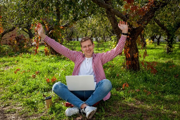 Facet siedzi na zielonej trawie w parku i pracuje na laptopie. Niezależna praca na zewnątrz. Praca poza biurem w związku z pandemią Covid 19