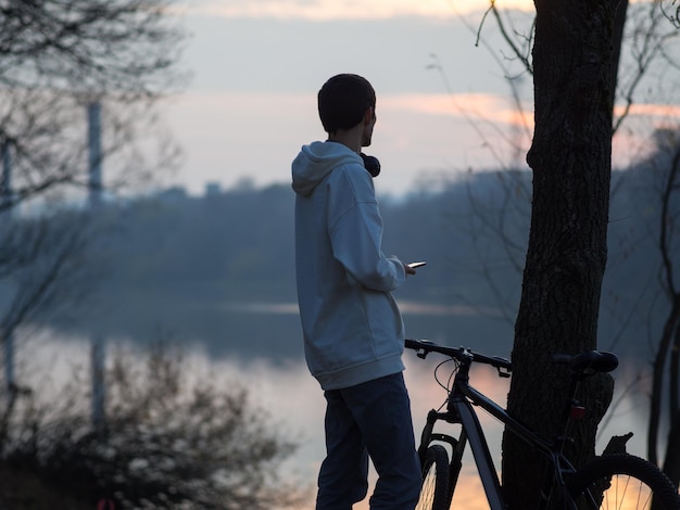Facet w białej bluzie z kapturem podziwia jesienny zachód słońca nad brzegiem rzeki i relaksuje słuchając muzyki przez słuchawki