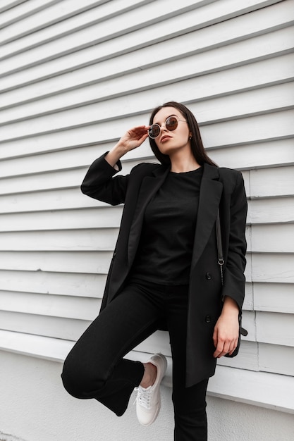 Fajna nowoczesna młoda kobieta w modnej czarnej odzieży casual z torebką prostuje okulary w pobliżu zabytkowego drewnianego budynku na ulicy w mieście. Amerykańska modna dziewczyna modelka odpoczywa na zewnątrz.