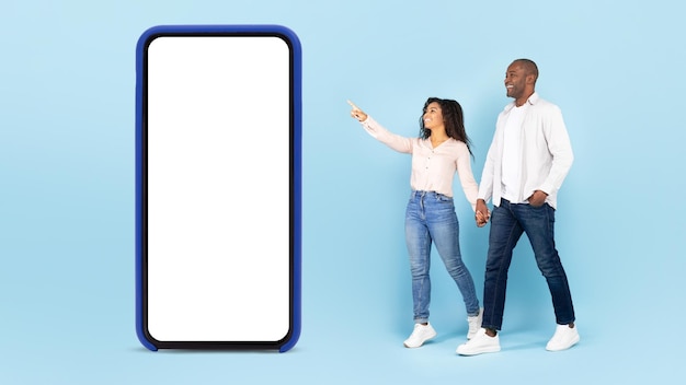 Fajna oferta Czarni małżonkowie spacerujący i dama wskazująca na duży smartfon z makietą miejsca na kopię promującą aplikację lub stronę internetową