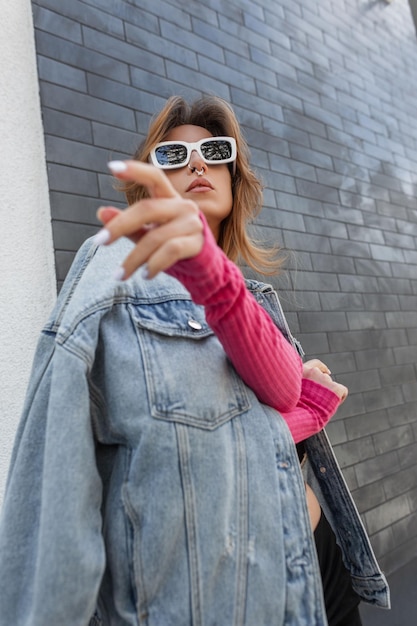 Fajna stylowa młoda kobieta hipster model z białymi fajnymi okularami przeciwsłonecznymi w modnym miejskim stroju z kurtką dżinsową i różową bluzą pozuje w pobliżu ceglanej czarnej ściany w mieście