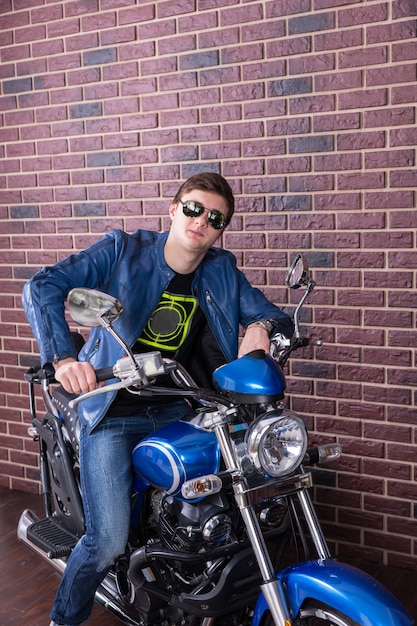 Fajny koleś siedzący na motocyklu w modnych, nowoczesnych okularach przeciwsłonecznych i skórzanej kurtce, oparty o kierownicę, patrzący w kamerę