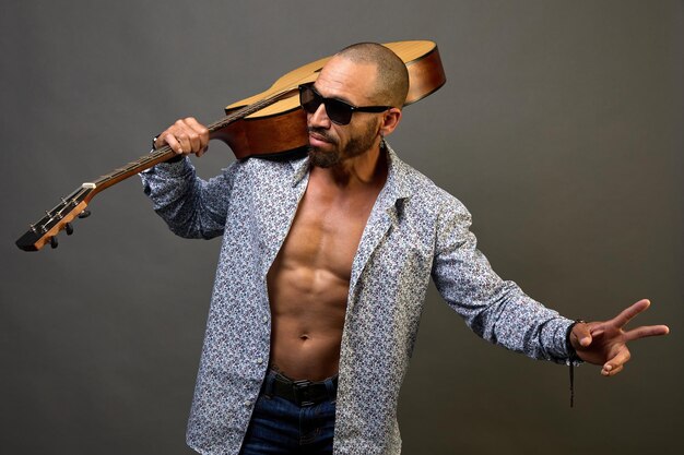 Fajny muskularny Latynos w okularach przeciwsłonecznych trzyma na ramieniu gitarę akustyczną