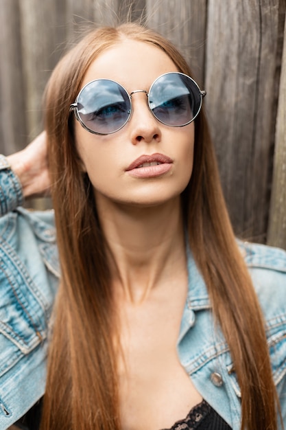 Fajny portret młodej kobiety w modnych niebieskich okularach przeciwsłonecznych w stylowej dżinsowej kurtce na zewnątrz
