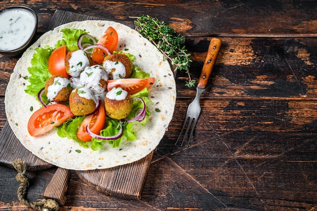 Falafel wegetariański z warzywami i sosem tzatziki na chlebie tortilla. Ciemne drewniane tło. Widok z góry. Skopiuj miejsce.