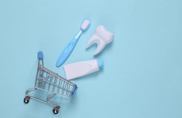 Fałszywy ząb, szczoteczka i pasta do zębów w wózku na zakupy na niebieskim tle. Widok z góry