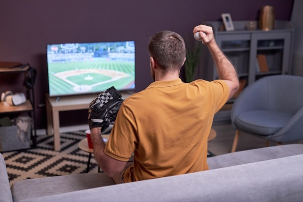 Fan baseballu ogląda mecz w telewizji w domu i trzyma piłkę baseballową