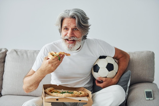 Fan piłki nożnej ogląda mecz i je pizzę Starszy stylowy współczesny mężczyzna z siwymi włosami i brodą w pomieszczeniu