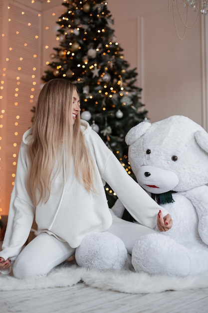 Fanny piękna szczęśliwa blondynka w fantazyjnej białej bluzie z kapturem i spodniami siedzi i cieszy się w pobliżu niedźwiedzia na podłodze w pobliżu ozdób choinkowych