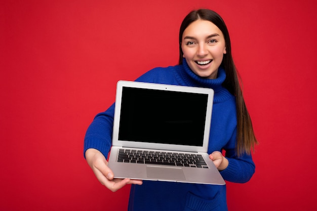 Fascynujący całkiem szczęśliwy piękna młoda brunet kobieta trzyma komputer laptop patrząc na kamery na sobie niebieski sweter na białym tle nad czerwonym tle ściany. Wolne miejsce na reklamę