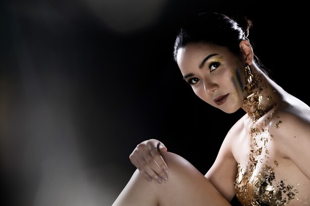 Fashion Young Asian Woman oczy owinięte czarnymi włosami piękny makijaż ozdobiony złotą folią lub złotym liściem na całej klatce piersiowej. Oświetlenie studyjne ciemne tło
