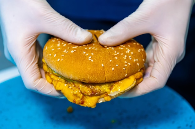 Fast food. Widok makro burgera. Ręce w białych lateksowych rękawiczkach trzymające burgera, pokazujące smaczny ser i kotlet. Zbliżenie