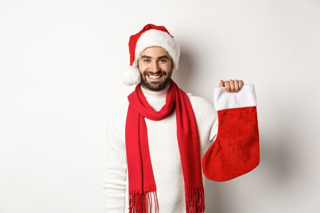 Ferie zimowe i koncepcja uroczystości. Szczęśliwy brodaty mężczyzna w santa hat trzymający świąteczną skarpetę, przynoszący prezenty, stojący na białym tle