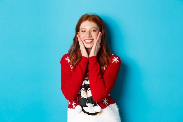 Ferie zimowe i koncepcja Wigilii. Wesoła i urocza ruda dziewczyna w świątecznym swetrze, rumieniąc się i dotykając policzków, uśmiechając się i radując na niebieskim tle.