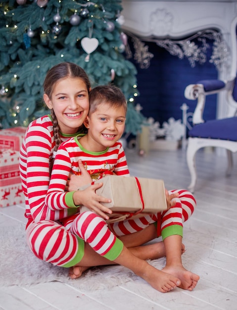 Figlarny uśmiechający się szczęśliwy ładny chłopak i dziewczyna ubrana w pasiastą piżamę z prezentem siedzi w urządzonym pokoju noworocznym w domu. Świąteczny dobry nastrój. Styl życia, rodzina i wakacje 2018 koncepcja