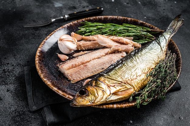 Filet śledziowy wędzony na gorąco na rustykalnym talerzu z tymiankiem i rozmarynem. Czarne tło. Widok z góry.