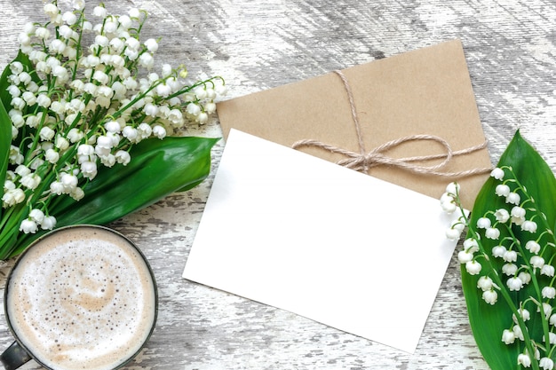 Filiżanka cappuccino z pustą białą kartkę z życzeniami i kopertę z wiosennymi kwiatami konwalii
