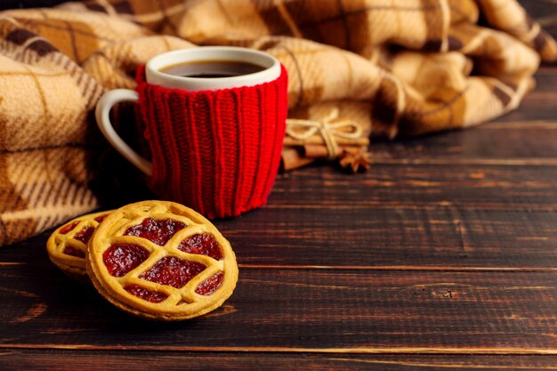 Filiżanka gorącej kawy, w dzianej okładce, domowych ciasteczkach i przyprawach, leży na drewnianym stole w pobliżu kraciastej kratki.