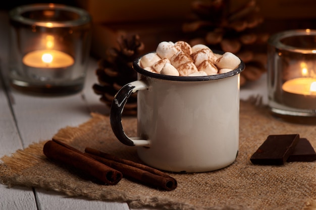 Filiżanka gorący kakao lub gorąca czekolada z piankami i cynamonowymi kijami na drewnianym tle z płonącymi świeczkami. Rustykalny. Zimowy nastrój.