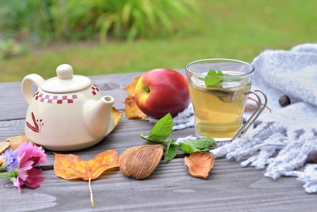 Filiżanka herbaty na drewnianym stole w ogrodzie z imbrykiem wśród jesiennych liści i czerwone jabłko na wełnianym szaliku