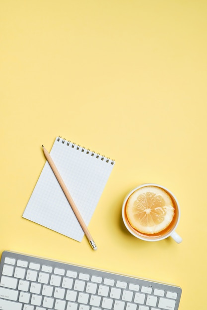 Filiżanka herbaty z cytryną obok klawiatury, notatnik i ołówek na żółtym tle. widok z góry.