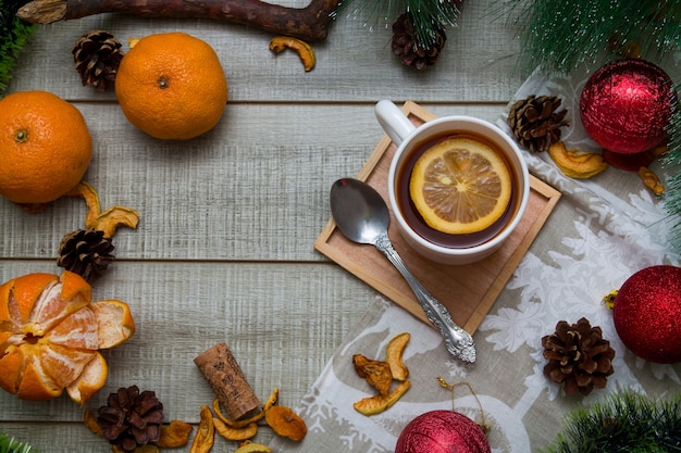Filiżanka herbaty z cytrynowym noworocznym wystrojem na drewnianym tle szary stół, zielona gałązka sosny, mandarynki, łyżka, suszone owoce, bombki, widok z góry, kopia przestrzeń