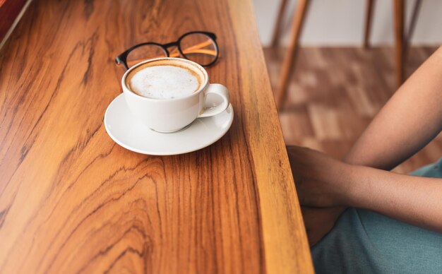 Filiżanka kawy cappuccino z latte i szklanki kobiety na drewnianym blacie barowym w nasłonecznionej kawiarni