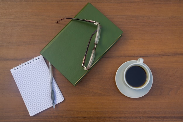 Filiżanka kawy, książka, notatnik, długopis i okulary na drewnianym biurku