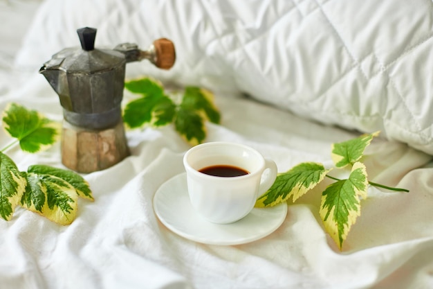 Filiżanka kawy na białej pościeli z ekspresem do kawy z gejzerem i zielonymi liśćmi
