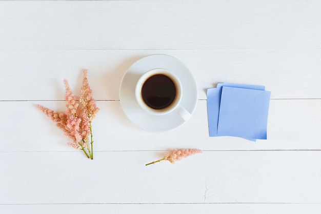 filiżanka kawy, różowe kwiaty i niebieskie papierowe naklejki na białym drewnianym stole. Widok z góry, Flatlay.