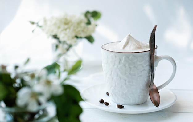 Zdjęcie filiżanka kawy z bitą śmietaną na białym drewnianym stole cappuccino w białej filiżance z łyżką modne poranne jasne kwiaty i ziarna kawy koncepcja dzień dobry