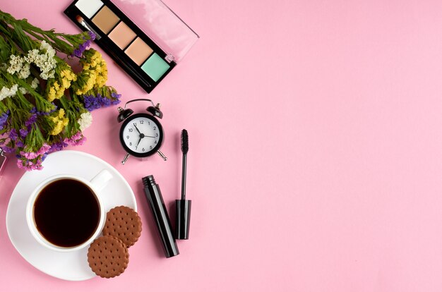 Filiżanka kawy z ciasteczkami, budzik, kwiaty, tusz do rzęs, na różowej powierzchni.