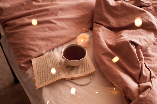 Filiżanka świeżej gorącej kawy pobyt na otwartej papierowej książce z płonącą świecą i świecące lampki świąteczne w zbliżenie łóżko. Zimowy sezon wakacyjny. Dzień dobry. Selektywne skupienie.