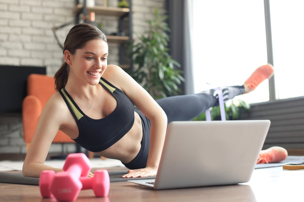 Fitness piękna szczupła kobieta robi deski boczne z taśmą oporową i ogląda samouczki online na laptopie, trening w salonie. Pozostań w domu.