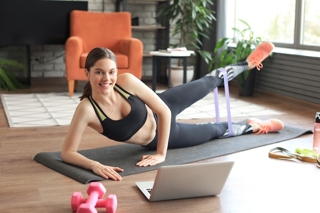 Fitness piękna szczupła kobieta robi deski boczne z taśmą oporową i ogląda samouczki online na laptopie, trening w salonie. Pozostań w domu.