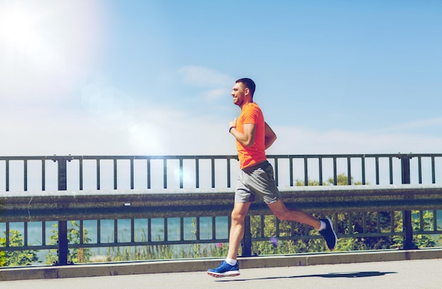 fitness, sport, ludzie, technologia i koncepcja zdrowego stylu życia - uśmiechnięty młody człowiek z zegarkiem tętna działa na letnim wybrzeżu