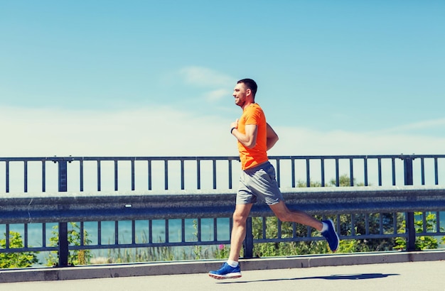 fitness, sport, ludzie, technologia i koncepcja zdrowego stylu życia - uśmiechnięty młody człowiek z zegarkiem tętna działa na letnim wybrzeżu