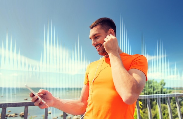 fitness, sport, ludzie, technologia i koncepcja zdrowego stylu życia - uśmiechnięty młody człowiek ze smartfonem i słuchawkami słuchający muzyki na letnim wybrzeżu nad falą dźwiękową lub diagramem sygnału