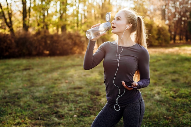 Fitness w parku, dziewczyna wody pitnej, trzymając w ręku smartfon i słuchawki.